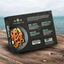 A box of The Spice Lab Seafood Seasoning Collection by The Spice Lab rests on a wooden surface beside the ocean, showcasing shrimp on a plate and highlighting seasoning blend descriptions like Shrimp and Crab Boil on its packaging.