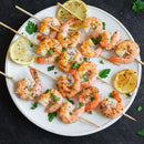 Grilled shrimp skewers garnished with herbs and lemon slices on a white plate, set on a dark surface, featuring bold flavors from The Spice Lab Seafood Seasoning Collection by The Spice Lab.