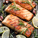 Grilled salmon fillets topped with fresh rosemary and a touch of The Spice Lab Seafood Seasoning Collection by The Spice Lab, surrounded by lemon slices, garlic cloves, and peppercorns on a grill pan.