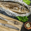 A whole baked fish, seasoned with cracked pepper and The Spice Lab Seafood Seasoning Collection, rests on a bed of lettuce in foil, accompanied by a brown bowl of herb butter, a beige napkin, and vintage fork and knife atop a dark surface.