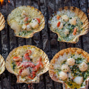 Six scallop shells filled with scallops and herbs are grilling over hot coals, bubbling with melted butter and garnished with chopped green herbs from The Spice Lab Seafood Seasoning Collection by The Spice Lab.
