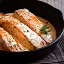 Three seared salmon fillets, perfectly seasoned with The Spice Lab Seafood Seasoning Collection, in a creamy herb sauce, garnished with fresh parsley, served in a black skillet on a striped cloth.