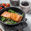 Grilled salmon fillet on a bed of fresh greens in a black skillet, garnished with tomatoes and served on a rustic wooden board, with marble salt and pepper containers in the background—a perfect dish to showcase flavors from The Spice Lab Seafood Seasoning Collection by The Spice Lab or your favorite Salmon Seasoning.