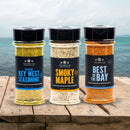 Three spice jars from The Spice Lab Seafood Seasoning Set—Key West Seasoning, Smoky Maple, and Best of the Bay—are displayed on a wooden surface with the ocean and sky softly blurred in the background, making them the perfect addition to any seafood seasoning collection by The Spice Lab.