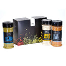 Three spice bottles from The Spice Lab Seafood Seasoning Set—featuring Classic Key West Seasoning, Smoky Maple, and Best of the Bay—ideal for seasoning salmon or adding flavor to seafood boils, are displayed in front of a decorative black gift box from The Spice Lab adorned with colorful herb and spice graphics.