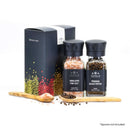 The Spice Lab Himalayan Pink Salt & Premium Black Pepper Grinder Set is displayed in front of a decorative box. The Himalayan pink salt jar and the black pepper grinder, both from The Spice Lab, are shown alongside wooden spoons holding salt and Tellicherry peppercorns. Spoons not included.