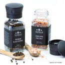 Two spice jars from The Spice Lab, labeled Himalayan Pink Salt and Premium Black Pepper, rest on a white surface alongside wooden spoons filled with Tellicherry Peppercorns and salt. Scattered salt crystals enhance the elegant display of The Spice Lab Himalayan Pink Salt & Premium Black Pepper Grinder Set.