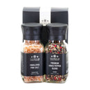 The Spice Lab Himalayan Pink Salt & Premium Kings Pepper Grinder Set features two glass grinders with black lids—one filled with pink Himalayan salt and the other with premium black pepper—displayed in front of a sleek, branded box from The Spice Lab, making it a perfect addition to any gourmet kitchen.