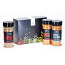 Three spice jars labeled Smoked Butchers Blend, Nashville Hot Chicken, and Spicy Seafood are arranged beside a decorative black box featuring a vibrant, seasoning-themed design. The Spice Lab Best Seller Seasoning Set by The Spice Lab offers an easy way to elevate your meals with bold grilling spices and rich hot chicken seasoning.