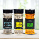 Three versatile blends from The Spice Lab Cooking Essentials Seasoning Set—butcher’s salt & pepper, lemon pepper, and chili lime—stand side by side on a wooden surface against a softly blurred background. This premium set from The Spice Lab brings together essential grilling spices perfect for any kitchen or cookout.