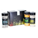 Three spice jars—butchers salt + pepper, chili lime seasoning, and lemon pepper—stand in front of a black box with vibrant spice graphics, creating The Spice Lab Cooking Essentials Seasoning Set by The Spice Lab: a perfect assortment of grilling spices and versatile seasonings for any kitchen.