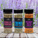 Three seasoning jars labeled Herbs de Provence, French Onion + Garlic, and Rosemary Roasted Garlic from The Spice Lab are displayed on a wooden surface with blooming lavender flowers in the background. The Spice Lab Flavors of Provence Seasoning Set is perfect for adding gourmet blends to your dishes and brings the essence of French and Italian-inspired cooking into your kitchen.