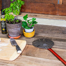 A kitchen scene featuring potted plants, a bottle of olive oil, a knife on a wooden cutting board, and the WPPO 7" Round Turning Pizza Peel with Break Down Handle by WPPO LLC Direct, distinguished by its red handle and resting on a rustic wooden table.