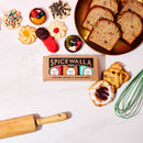 A gold plate showcases an array of assorted cookies and Pumpkin Spice quick bread slices, accompanied by the Spicewalla 3 Pack Holiday Baking Collection, a rolling pin, a whisk, and a pastry, all artfully arranged on a white surface.
