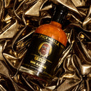 A bottle of Spicewalla T-Pain x Spicewalla - The Sauce (Perfectly Imperfect Edition) rests on gleaming, crumpled gold fabric, providing a luxurious backdrop for the dark-labeled sweet and smoky hot sauce from Spicewalla.