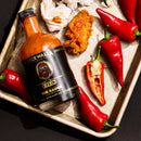A metal tray featuring red chili peppers, a halved pepper, a bottle of Spicewalla T-Pain x Spicewalla - The Sauce (Perfectly Imperfect Edition) hot sauce, a piece of fried chicken, and pieces of crumpled paper arranged on a dark surface.