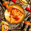 A slice of cheese pizza topped with sauce and red peppers rests on a gold plate, surrounded by tacos, Spicewalla T-Pain x Spicewalla – The Sauce (Perfectly Imperfect Edition) bottles, and red peppers on a metallic brown fabric background—the perfect setting for a Spicewalla Perfectly Imperfect Edition feast.