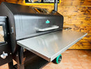 A Pimp My Grill® Stainless Steel Shelf Extension / Sleeve for Green Mountain Grills Jim Bowie / Peak attached to the front of a black pellet grill.