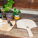 A WPPO LLC Direct 12" x 24" Wooden Pizza Peel (Launch Pad) from the 2 Pack rests alongside potted plants, olive oil, a knife, and dough on a rustic wooden countertop in a cozy kitchen setting.