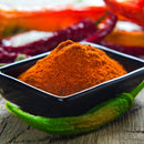 A black square bowl filled with vibrant orange-red The Spice Lab Ground Cayenne Pepper sits on a rustic wooden surface, with whole red and green chile peppers placed nearby.