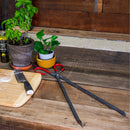 Displayed on a rustic wooden table are WPPO Forged Steel Wood Pliers from WPPO LLC Direct, red-handled scissors, a kitchen knife, potted plants, and a bottle of olive oil—ideal companions for prepping ingredients beside your outdoor or wood-fired oven.