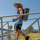 A man in a straw hat, sunglasses, blue tank top, and shorts walks on a dock ramp by a lake, carrying the Bob The Cooler Co. 25L "The Bro" Backpack Soft Cooler from Bob - The Cooler Co. and holding a blue mug.