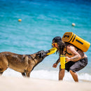 A man and his dog play tug-of-war with a yellow rescue float on a sandy beach by the ocean. The man, wearing sunglasses and a yellow flotation backpack, keeps his Bob The Cooler Co. 25L "The Bro" Backpack Soft Cooler by his side, ready to enjoy chilled drinks under the sun.