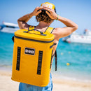 A person wearing a yellow cap and carrying the Bob The Cooler Co. 25L "The Bro" Backpack Soft Cooler by Bob - The Cooler Co. stands on a beach facing the sea, with boats visible in the blue water under a clear sky.