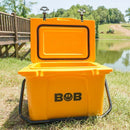 A bright yellow Bob The Cooler Co. 25QT / 23.6L "Wingman" Hard Cooler by Bob - The Cooler Co. sits open on the grass near a pond, with trees and a wooden bridge in the background.