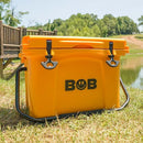 A vibrant yellow Bob The Cooler Co. 25QT / 23.6L "Wingman" Hard Cooler featuring black latches rests on the grass beside a pond, with a wooden bridge and trees in the background beneath a clear sky. This durable, commercial-grade insulated cooler from Bob - The Cooler Co. is perfect for keeping your drinks cold while you unwind outdoors.