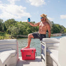 A shirtless man with long blonde hair and sunglasses poses on a boat, holding up a drink. He has one foot resting on a red Bob The Cooler Co. 25QT / 23.6L "Wingman" Hard Cooler by Bob - The Cooler Co., featuring a convenient bottle opener, surrounded by water and trees beneath a partly cloudy sky.