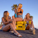 Three women in summer outfits sit on a rooftop with drinks, smiling and chatting. Next to them is the Bob The Cooler Co. 25QT/23.6L "Wingman" Hard Cooler by Bob – The Cooler Co., its bright yellow exterior ensuring their refreshments stay perfectly chilled. Palm trees and a clear sky create a relaxed backdrop.