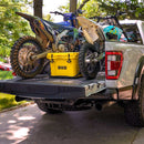 A pickup truck tailgate is open with two dirt bikes loaded in the bed. A yellow Bob The Cooler Co. 25QT / 23.6L “Wingman” Hard Cooler from Bob - The Cooler Co. sits between the bikes. Trees and grass are visible in the background.