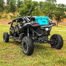 A black off-road vehicle with oversized tires is parked on grass. Two fishing rods are mounted at the back, and a blue Bob The Cooler Co. 25QT "Wingman" hard cooler from Bob - The Cooler Co. is strapped to the rear. Trees and hills can be seen in the background.