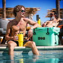 Two men relax by the pool; one sits on the edge wearing sunglasses and holding a yellow tumbler and cooler. The other lounges behind him. Beside them, under a sunny sky, is a Bob - The Cooler Co. 25QT / 23.6L "Wingman" Hard Cooler labeled “BOB.”.