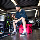 A man in casual clothes and a backwards cap sits on the Bob The Cooler Co. 25QT / 23.6L "Wingman" Hard Cooler in a garage, looking at his phone and holding a water bottle. Tools, motorcycle gear, and the handy built-in bottle opener from Bob - The Cooler Co. can be seen in the background.