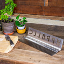 A metal chopping guide with "CHOPPER" cut out sits on a wooden table next to a cutting board with a knife, flour, and potted plants. This setup is ideal for any kitchen enthusiast, especially for those aiming to elevate their pizza-making experience using the WPPO Wood-Fired Oven Flame / Heat Deflector by WPPO LLC Direct, a popular stainless steel pizza oven accessory designed to ensure even cooking in wood-fired ovens.