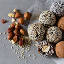 Assorted energy balls coated with The Spice Lab Organic Hulled Sesame Seeds, shredded coconut, and cocoa powder are arranged on parchment paper next to a pile of hazelnuts and scattered sesame seeds—perfect for baking or adding a nutritious touch to your favorite stir-fries.