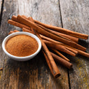 A small white bowl filled with The Spice Lab Organic Ground Vietnamese Saigon Cinnamon sits on a rustic wooden surface, surrounded by several whole high-oil cinnamon sticks from The Spice Lab.