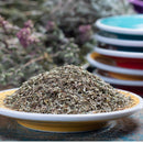 A close-up of a small plate filled with The Spice Lab Savory Whole Leaf 1 lb, with a blurred background featuring stacked colorful dishes and additional foodservice herbs.