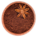 A wooden bowl filled with The Spice Lab Ground Star Anise, topped with a whole star anise on one side, is seen from above on a white background. This aromatic spice from The Spice Lab adds a sweet licorice-like flavor to enhance your favorite recipes.