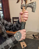 A person in a plaid shirt holds the Custom Handmade Viking Axe by Texan Knives, featuring high carbon steel, intricate engravings, and a leather-wrapped handle. Their hand displays a Norse-style geometric tattoo.