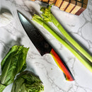 The Spaceman Knives EM Wave Chef Knife by Spaceman Knives, featuring a multicolored handle, sits on a marble countertop with celery, lettuce, garlic, and a wooden cutting board—a standout in any professional kitchen.