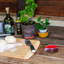 Two dough balls rest on a floured cutting board accompanied by a knife, olive oil, fresh herb plants, an herb grinder, and the WPPO Pizza Cutter With Removable Safety Shield by WPPO LLC Direct—its precision roller blade with detachable shield neatly displayed alongside the other items atop the wooden counter.