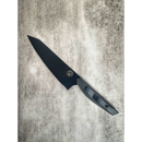 The Spaceman Knives Dark Matter Kiritsuke chef’s knife, made with premium 7CR17MOV-X steel, rests diagonally on a textured gray surface, its sleek black blade and handle angled toward the upper left corner.