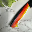 A close-up of the Spaceman Knives EM Wave Chef Knife handle, featuring a vibrant red, orange, yellow, and green gradient, rests on white marble with leafy greens—showcasing Spaceman Knives’ blend of color and professional kitchen elegance.