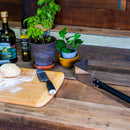 A wooden table set with dough on a cutting board, a knife, a spatula, potted plants, a bottle of olive oil, a spice grinder, and the WPPO Coal Rake for Wood-Fired Pizza Oven from WPPO LLC Direct—everything you need to prepare fresh ingredients before baking in your wood-fired pizza oven.