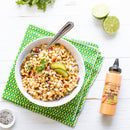 A bowl of corn salad with avocado and cilantro sits on a colorful napkin, alongside sliced limes, a metal spoon, pepper, and Terrapin Ridge Farms Chile Lime Aioli Squeeze Garnishing Sauce on a white wooden table.