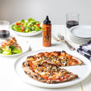 A table set with a vegetable pizza topped with Terrapin Ridge Farms Truffle Hot Sauce, salad, two glasses of red wine, a bottle of Terrapin Ridge Farms gourmet condiment, stacked plates, forks, and a checkered napkin on a white surface.