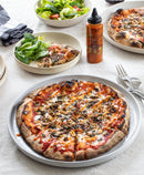 A pizza with mushrooms and Terrapin Ridge Farms Truffle Hot Sauce sits on a plate by forks. Nearby, a salad bowl, a small plate with salad and pizza slice, and a bottle of Terrapin Ridge Farms’ gourmet hot sauce rest on a white tablecloth.